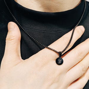 May include: A black necklace with a small, round pendant featuring a stylized letter. The chain is a delicate, linked design. The pendant hangs from the chain, resting on a black shirt.