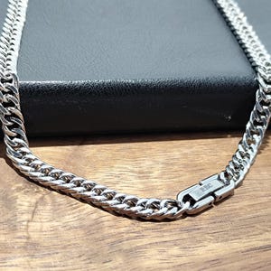 May include: A silver-colored curb chain necklace with a secure clasp. The necklace is displayed on a dark surface, with a wooden background. The chain links are closely interlocked, creating a bold and stylish accessory.