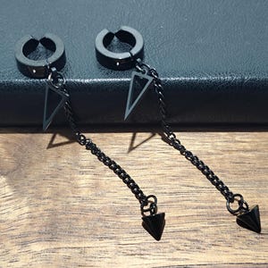 May include: Two black, non-piercing hoop earrings with dangling chains. Each earring features a triangular charm and a pointed, cone-shaped pendant. The earrings are displayed on a wooden surface with a dark background.