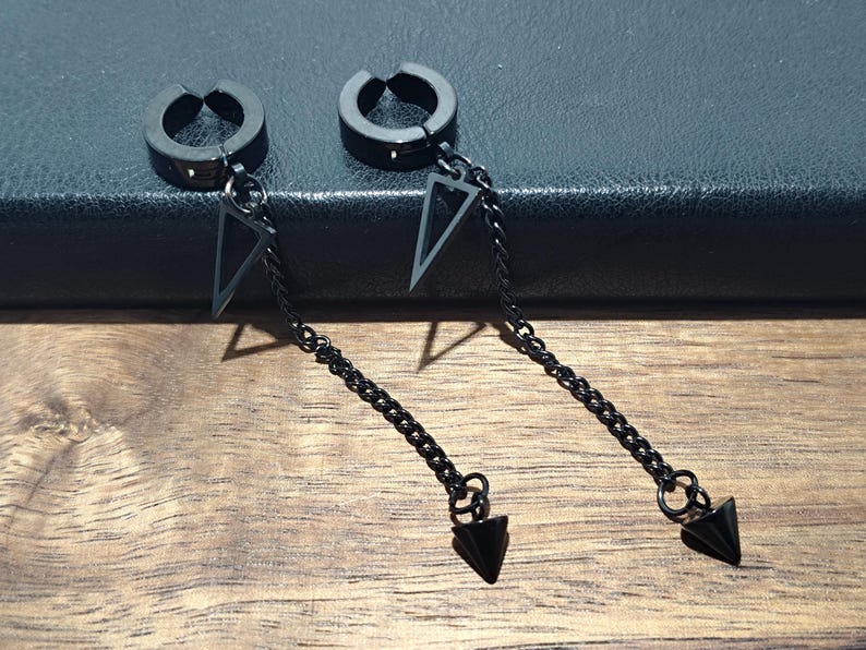 May include: Two black, non-piercing hoop earrings with dangling chains. Each earring features a triangular charm and a pointed, cone-shaped pendant. The earrings are displayed on a wooden surface with a dark background.