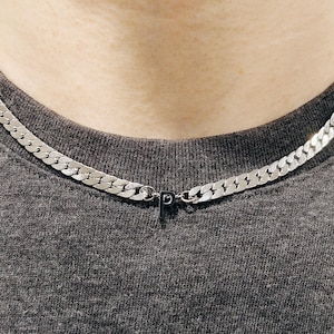 May include: A silver curb chain necklace with a small pendant featuring the letter "P". The necklace is worn around the neck over a gray t-shirt. The chain links are flat and closely connected.