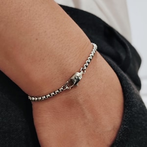 May include: A silver-colored bracelet with a rounded chain design. The bracelet is secured with a lobster clasp. The jewelry is worn on a wrist, with a dark background.