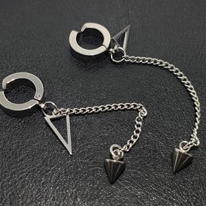May include: A pair of silver-toned ear cuffs with a chain and triangle and cone-shaped charms. The ear cuffs have a circular design that clips onto the ear. The chain connects the ear cuff to a triangle charm and a cone-shaped charm.