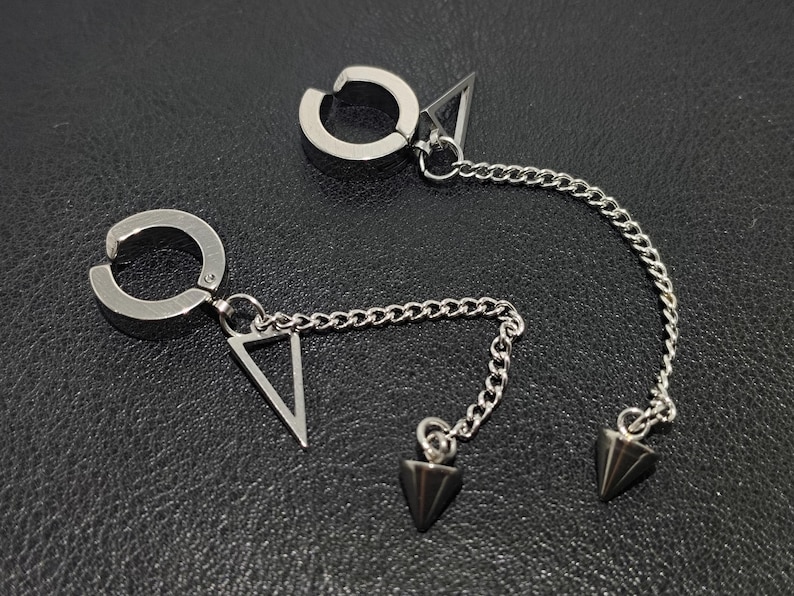May include: A pair of silver-toned ear cuffs with a chain and triangle and cone-shaped charms. The ear cuffs have a circular design that clips onto the ear. The chain connects the ear cuff to a triangle charm and a cone-shaped charm.