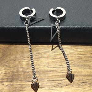 May include: A pair of silver-toned clip-on earrings. Each earring features a circular clip, a triangular charm, a chain, and a cone-shaped pendant. The earrings are displayed against a dark background.