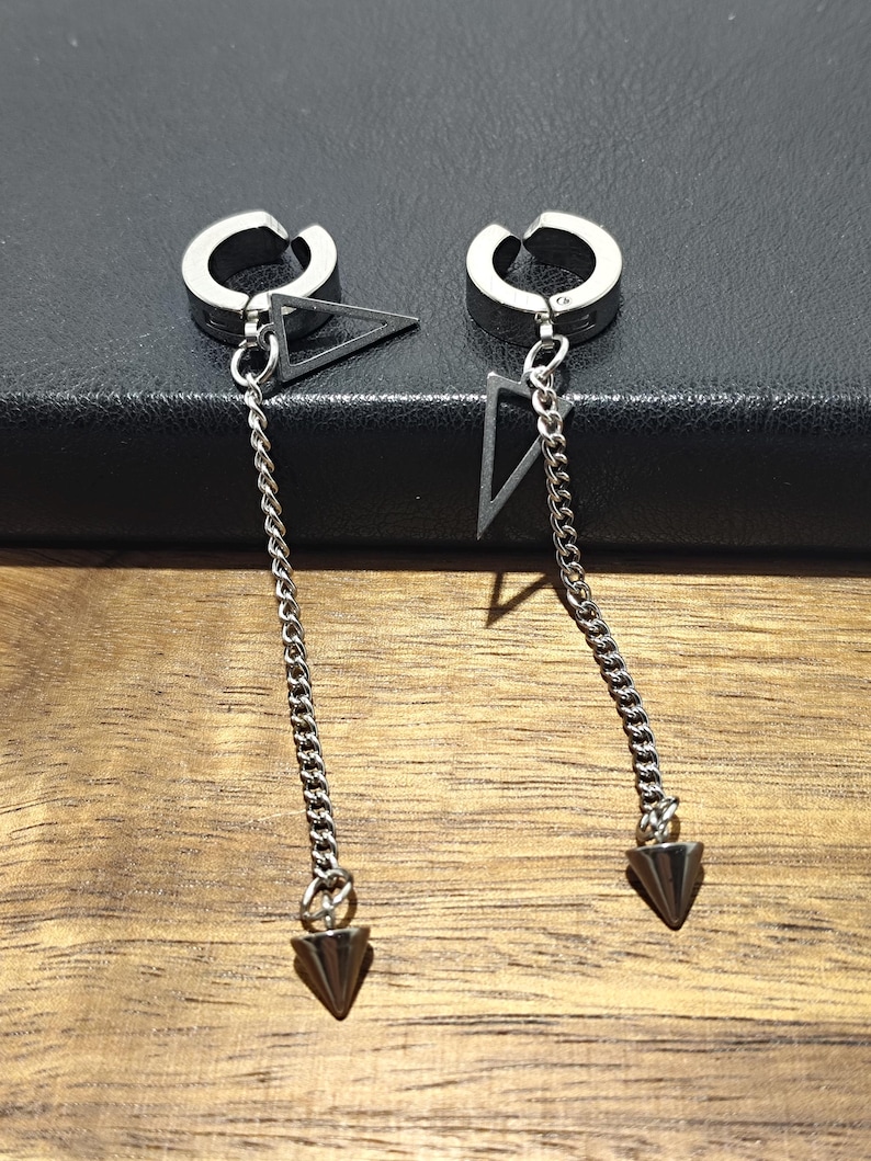 May include: A pair of silver-toned clip-on earrings. Each earring features a circular clip, a triangular charm, a chain, and a cone-shaped pendant. The earrings are displayed against a dark background.