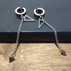 May include: A pair of silver-toned earrings. Each earring features a hoop that clips onto the ear, a triangle, a chain, and a cone-shaped charm. The earrings are displayed on a dark surface.