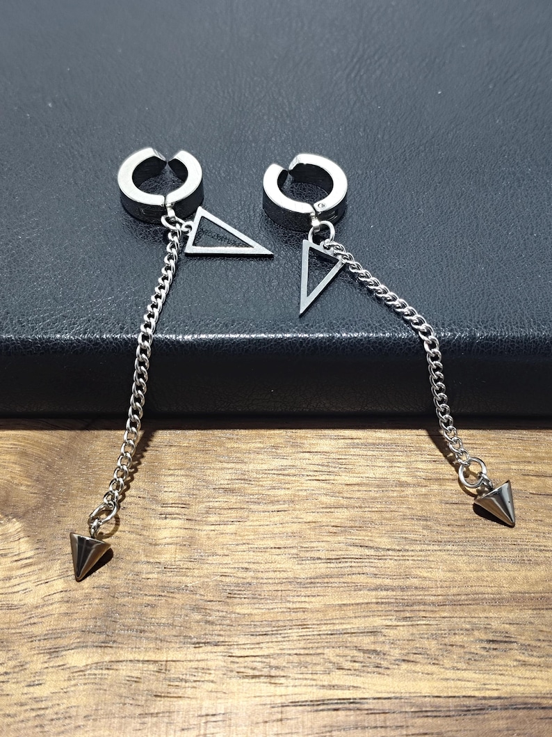May include: A pair of silver-toned earrings. Each earring features a hoop that clips onto the ear, a triangle, a chain, and a cone-shaped charm. The earrings are displayed on a dark surface.