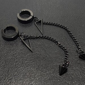 May include: A pair of black, non-piercing earrings. Each earring features a circular clip-on design with a dangling chain. The chain is attached to a triangular pendant and terminates in a pointed cone. The earrings are displayed on a textured, dark surface.