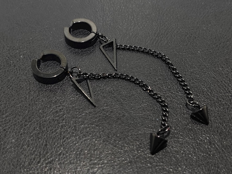 May include: A pair of black, non-piercing earrings. Each earring features a circular clip-on design with a dangling chain. The chain is attached to a triangular pendant and terminates in a pointed cone. The earrings are displayed on a textured, dark surface.