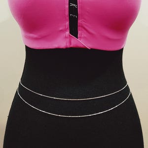 May include: A silver body chain with two layers. The chain is worn over a black top.