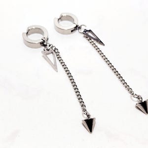 May include: A pair of silver-toned clip-on earrings. Each earring features a circular clip, a triangular charm, a chain, and a downward-pointing triangular pendant. The earrings are displayed on a white surface.