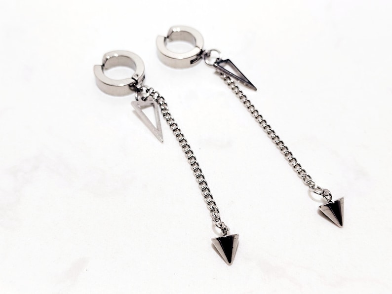 May include: A pair of silver-toned clip-on earrings. Each earring features a circular clip, a triangular charm, a chain, and a downward-pointing triangular pendant. The earrings are displayed on a white surface.