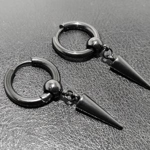 May include: A pair of black hoop earrings with a dangling, pointed cone design. The earrings have a small ball accent and are made of a dark, metallic material. The earrings are displayed on a textured, dark gray surface.