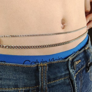 May include: A silver chain belt worn around the waist. The chain is layered in two rows and is secured with a clasp. The belt is worn over a blue waistband and blue jeans.