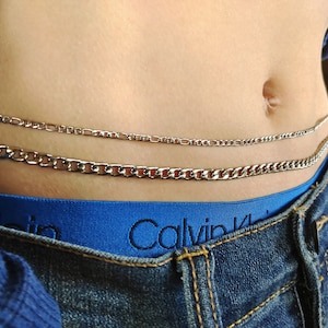 May include: Three silver chains layered around a person's waist. The chains are different widths and styles. The person is wearing blue jeans and a blue shirt.