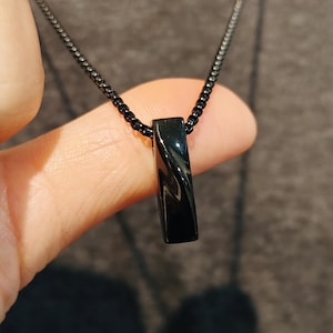 May include: A black pendant necklace featuring a rectangular, glossy pendant with a subtle twist design. The pendant hangs from a matching black chain. The necklace is held up by a person's fingers.