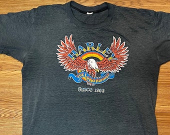 May include: Dark gray t-shirt featuring a colorful graphic of an eagle with outstretched wings, a rainbow, and the word "HARLEY". Below the eagle is the text "Since 1903". The shirt is laid flat on a wooden surface.