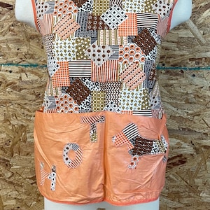 Puede incluir: Un delantal vintage de patchwork naranja y marrón con un bolsillo grande y la palabra "HOT" cosida en la parte delantera.
