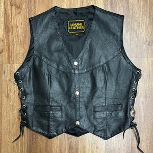 Może przedstawiać: Czarna skórzana kamizelka z sznurowaniem po bokach. Kamizelka ma spiczasty dół i metkę z napisem "GENUINE LEATHER".