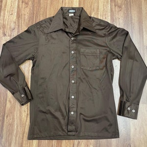May include: A brown long-sleeve button-up shirt with a single pocket on the left chest. The shirt has a collared neckline and a La Brique brand label.