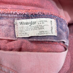 May include: Close-up of a pair of pink Wrangler jeans. The label reads "Wrangler" and includes washing instructions and size details. The jeans are made of 100% cotton.