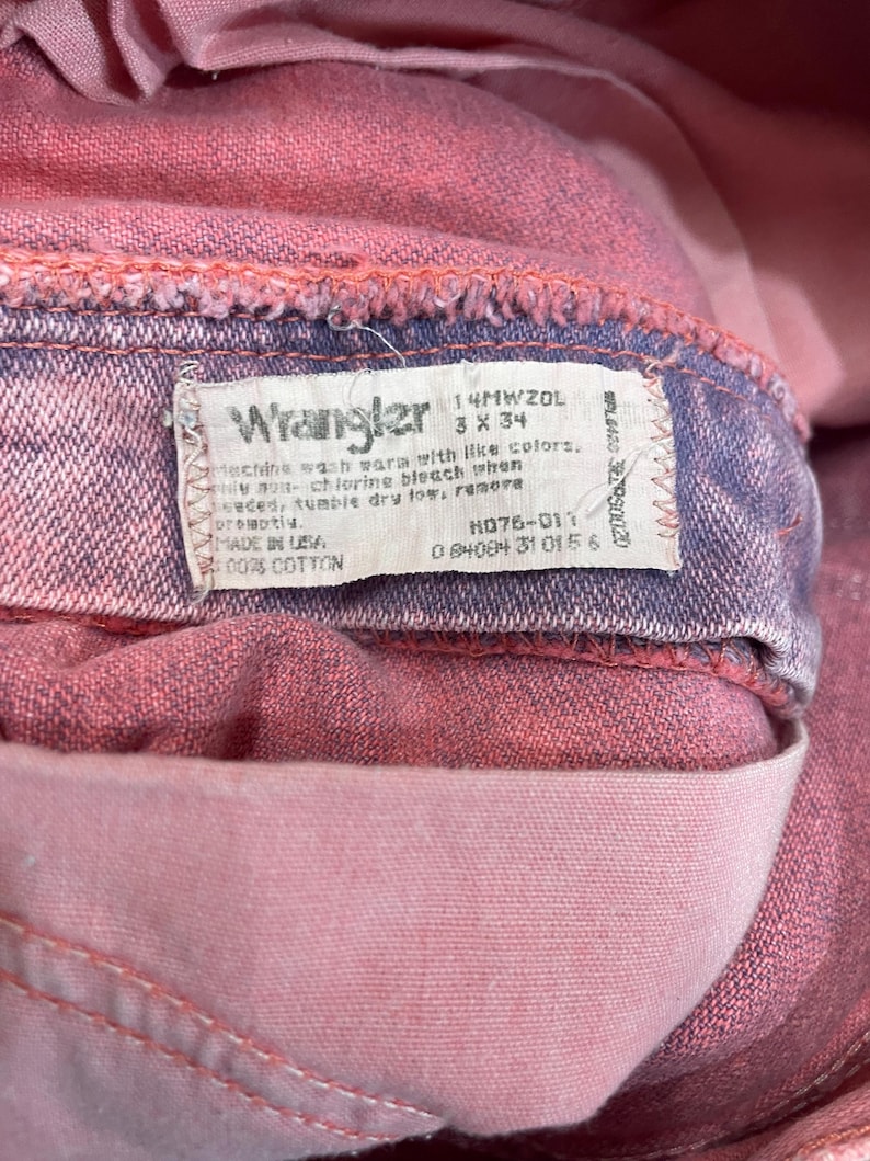 May include: Close-up of a pair of pink Wrangler jeans. The label reads "Wrangler" and includes washing instructions and size details. The jeans are made of 100% cotton.