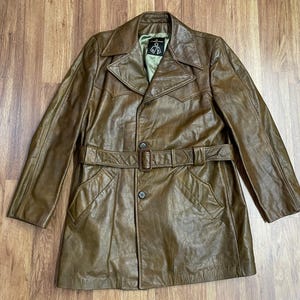 May include: A brown leather trench coat with a belt and a button closure. The coat has a collar and two pockets.