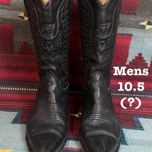 Op de afbeelding: Een paar zwarte leren cowboylaarzen met decoratieve stiksels en een puntige neus. De laarzen hebben maat 10,5. De laarzen worden gepresenteerd op een kleurrijk patroonkleed.