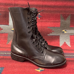 May include: Black leather combat boots with a lace-up design. The boots are mid-calf length and have a robust sole. The boots are displayed on a red patterned rug.