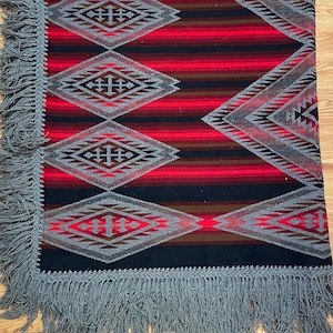 May include: A rectangular blanket with a geometric pattern in red, black, and gray. The design features diamond shapes and horizontal stripes. The blanket has a gray fringe along the edges, adding a decorative touch.