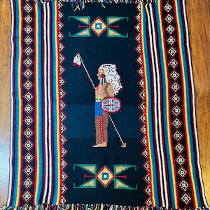 May include: A black woven throw blanket with a colourful striped border and a central image of a Native American warrior holding a spear and shield. The warrior is wearing a feathered headdress and a blue and white patterned shirt. The blanket has a fringe border.