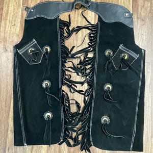 以下が含まれることがあります： 黒いスエードのチャップス。革製のウエストバンドと装飾的なシルバーのコンチョが特徴です。チャップスは前面とポケットにフリンジのディテールがあり、白いステッチが施されています。クラシックなウエスタンウェアアイテム。