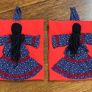 May include: Two red fabric pouches with a black yarn doll sewn on the front. The doll is wearing a blue and white floral dress with a red trim.