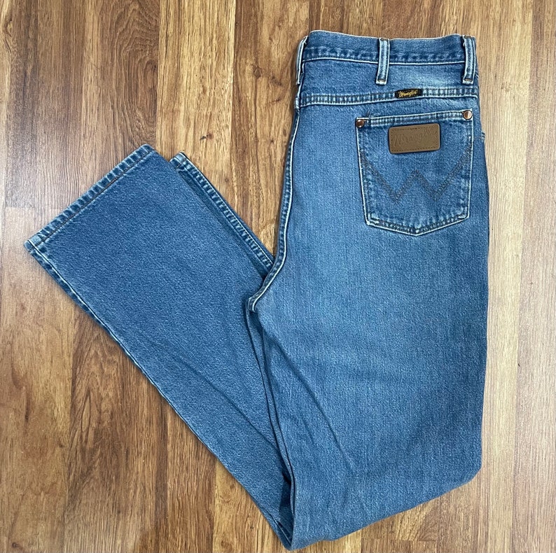 Może przedstawiać: Dżinsy z jasnoniebieskiego denimu z brązową sk&oacute;rzaną naszywką na tylnej kieszeni z napisem "Wrangler".