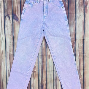 Pu&ograve; includere: Jeans viola chiaro a vita alta con un classico design a cinque tasche. I jeans hanno un aspetto sbiadito e un taglio dritto. Un'etichetta vintage &egrave; attaccata alla cintura. I jeans sono esposti su una superficie di legno.
