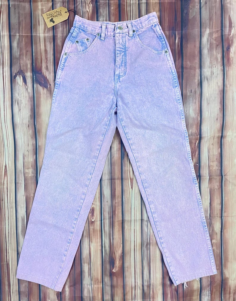 Pu&ograve; includere: Jeans viola chiaro a vita alta con un classico design a cinque tasche. I jeans hanno un aspetto sbiadito e un taglio dritto. Un'etichetta vintage &egrave; attaccata alla cintura. I jeans sono esposti su una superficie di legno.