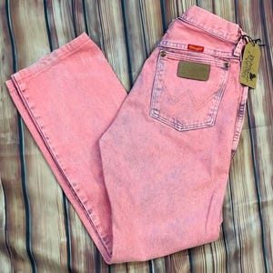 May include: Pink Wrangler jeans with a faded, vintage look. The jeans have a classic five-pocket design, a brown leather patch, and the Wrangler logo. A black tag with text hangs from the jeans.