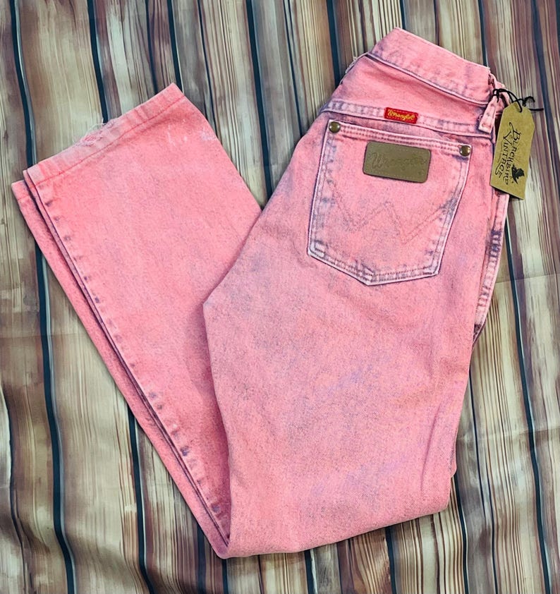 May include: Pink Wrangler jeans with a faded, vintage look. The jeans have a classic five-pocket design, a brown leather patch, and the Wrangler logo. A black tag with text hangs from the jeans.
