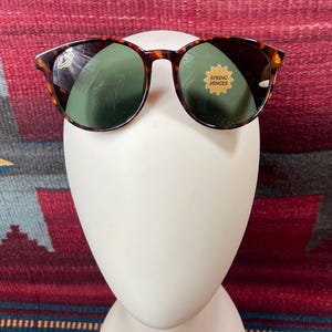 May include: Round sunglasses with a tortoiseshell frame and green lenses. The sunglasses have a sticker that reads "Spring Hinges". The glasses are displayed on a white mannequin head.