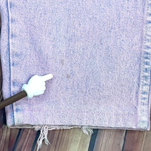 Pu&ograve; includere: Primo piano di jeans in denim viola chiaro con orlo strappato. I jeans hanno un aspetto sbiadito e mostrano segni di usura. Un puntatore bianco a forma di mano si trova nell'angolo in basso a sinistra, indicando una macchia sui jeans.