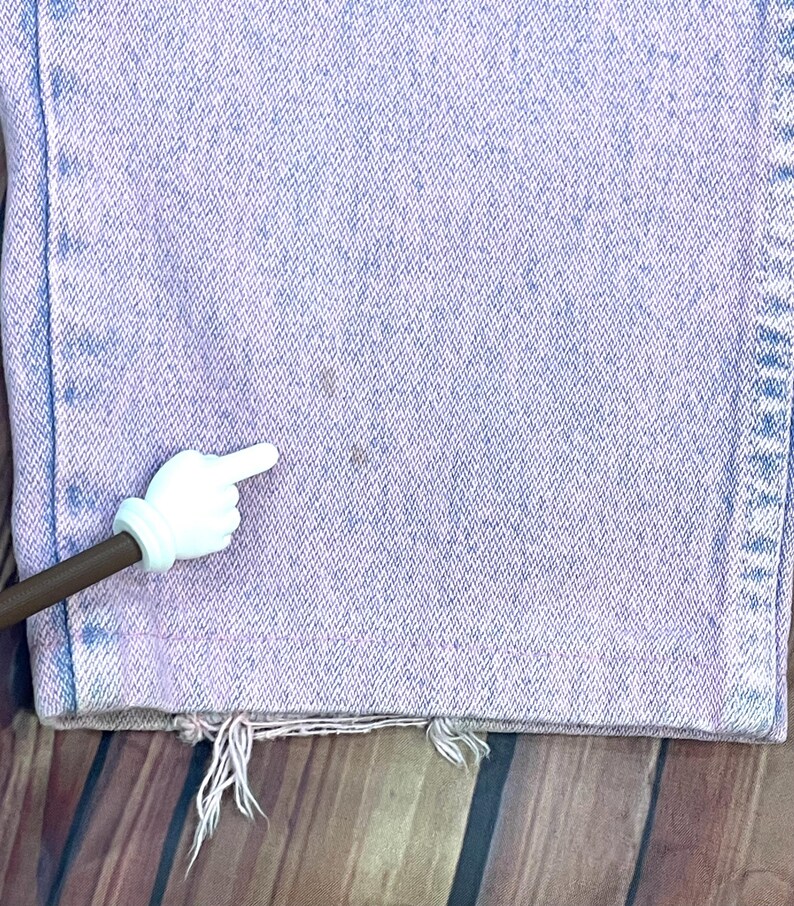 Pu&ograve; includere: Primo piano di jeans in denim viola chiaro con orlo strappato. I jeans hanno un aspetto sbiadito e mostrano segni di usura. Un puntatore bianco a forma di mano si trova nell'angolo in basso a sinistra, indicando una macchia sui jeans.