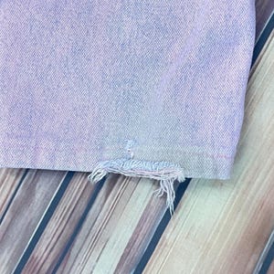 Pu&ograve; includere: Primo piano di una sezione di jeans in denim viola chiaro. L'orlo &egrave; sfilacciato, con fili sciolti. Il denim ha un aspetto leggermente sbiadito. Lo sfondo &egrave; una superficie in legno con assi diagonali.