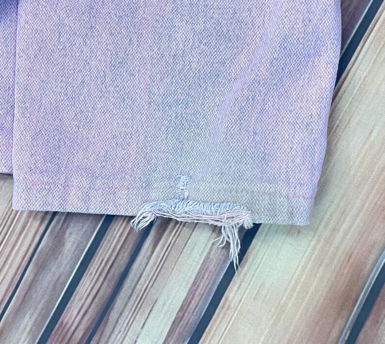 Pu&ograve; includere: Primo piano di una sezione di jeans in denim viola chiaro. L'orlo &egrave; sfilacciato, con fili sciolti. Il denim ha un aspetto leggermente sbiadito. Lo sfondo &egrave; una superficie in legno con assi diagonali.