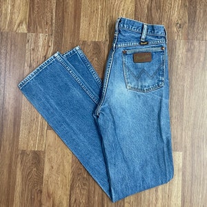 Può includere: Un paio di jeans blu denim con una toppa in pelle marrone Wrangler sulla tasca posteriore.