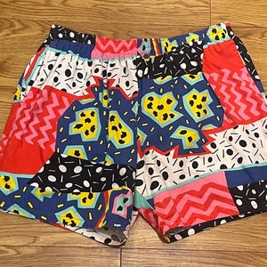 Könnte beinhalten: Bunte Shorts mit Muster im Patchwork-Design. Die Shorts zeigen eine Mischung aus roten, blauen, rosa, schwarzen und weißen geometrischen Formen, darunter Zickzackmuster, Punkte und abstrakte Muster. Der elastische Bund sorgt für Komfort.