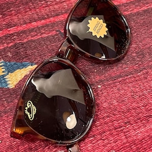 Könnte beinhalten: Sonnenbrille mit Schildpattmuster und dunklen Gläsern. Die Sonnenbrille hat eine runde Form und Federscharniere. Ein goldener Aufkleber auf dem rechten Glas trägt die Aufschrift "SPRING HINGES". Das linke Glas hat ein goldenes Logo und den Text "SS600".