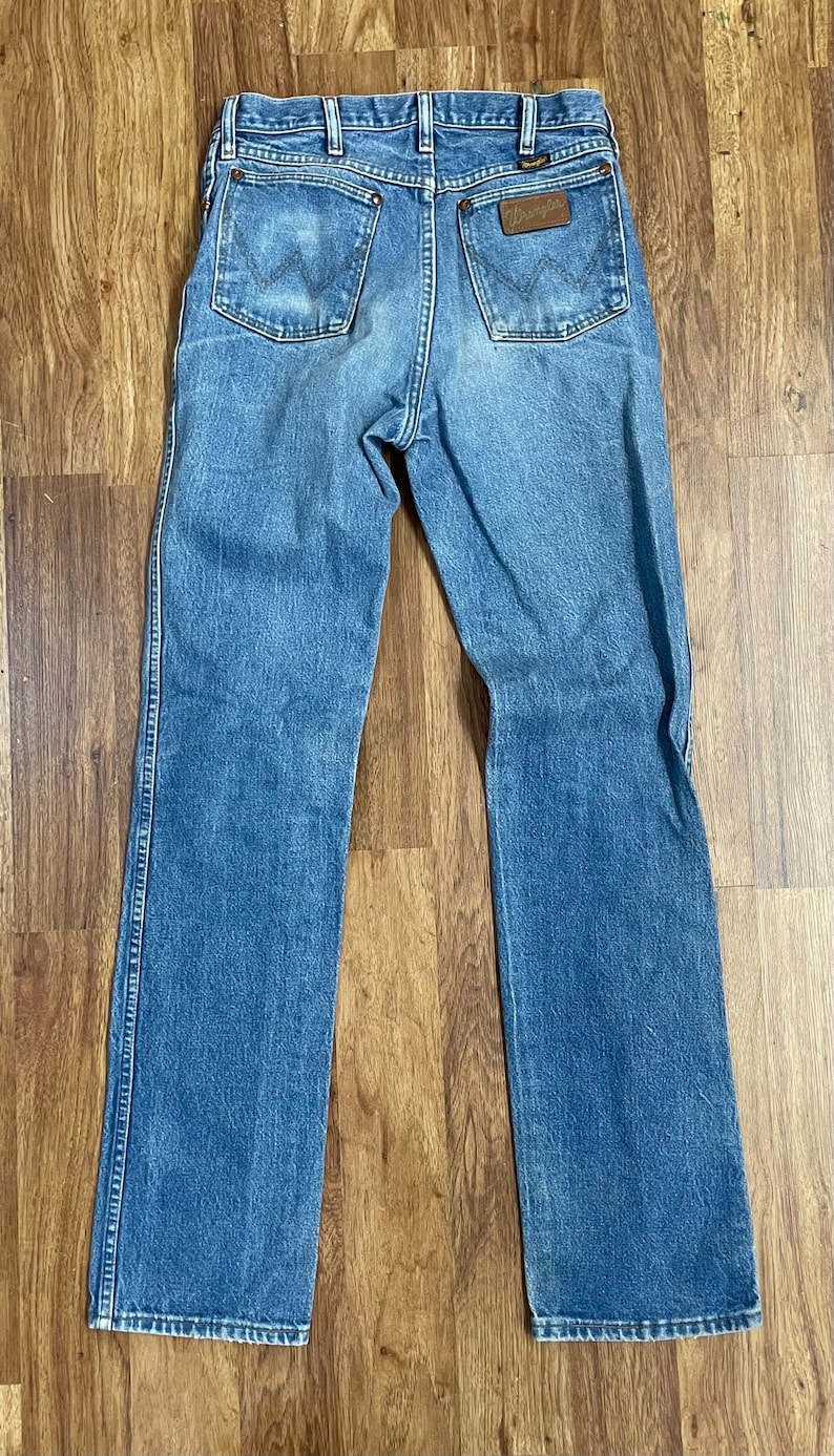 May include: A pair of blue denim jeans with a brown leather Wrangler label on the back pocket.