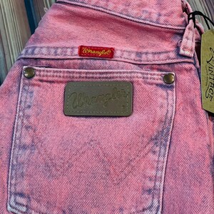 May include: Close-up of pink Wrangler jeans with a faded, vintage look. The back pocket features a brown leather patch with the Wrangler logo. A red Wrangler label is visible above the pocket. A vintage tag hangs from the jeans.
