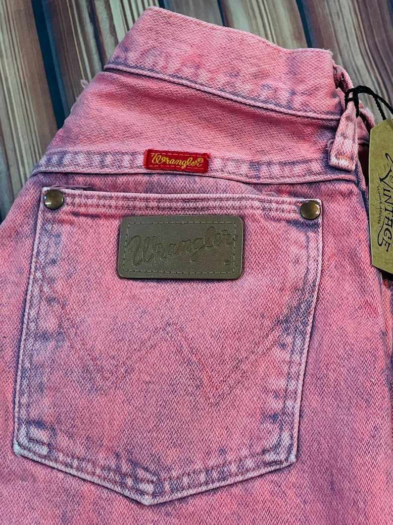 May include: Close-up of pink Wrangler jeans with a faded, vintage look. The back pocket features a brown leather patch with the Wrangler logo. A red Wrangler label is visible above the pocket. A vintage tag hangs from the jeans.